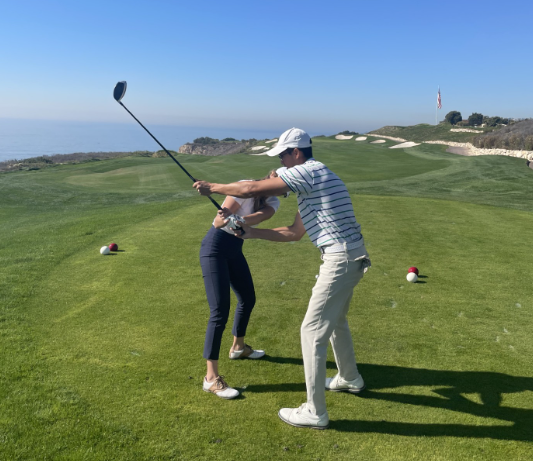 A man in a white hat coaching a female golfer on a green grass tee box.